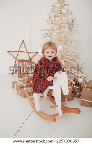 Christmas photo of a one-year-old girl in a red dress sitting on a rocking horse. Christmas photo near the tree. children's red checkered dress. children's Christmas room, wooden Christmas tree