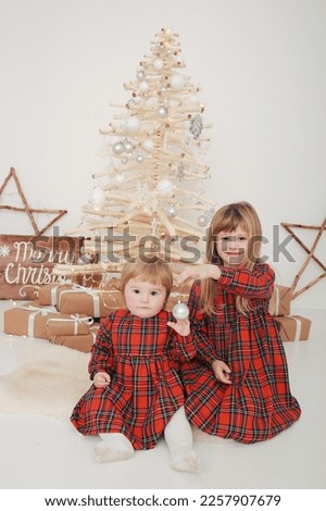 family Christmas two sisters are sitting near a wooden Christmas tree in red clothes. red plaid dress for Christmas. New Year's decor in the photo studio