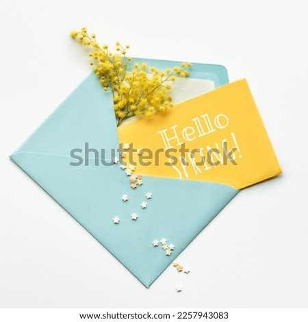 Caption Hello Spring on yellow paper card, fresh mimosa flowers in mint blue envelope on off white background. Natural springtime decor in two colors. Flat lay, top view, central composition. Royalty-Free Stock Photo #2257943083