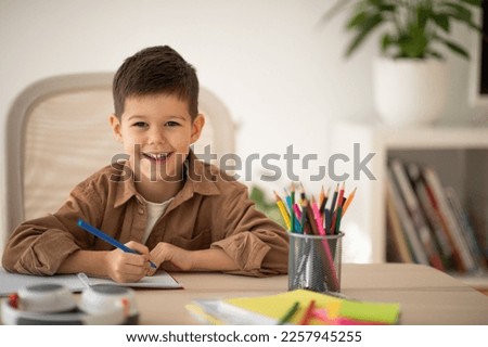 Smiling cute small european boy sitting at table, draws picture, look at camera in school, children garden room interior. Elementary education, art, study, learning at home and childhood lifestyle