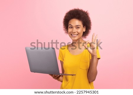 Glad millennial multiethnic woman in casual typing on computer show ok sign with hand isolated on pink background, studio. Recommendation to device for study, chat in social networks, ad and offer