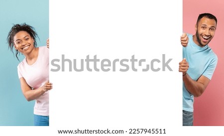 Promotion billboard. Excited black guy and lady holding white advertisement board, standing on halved pink and blue background, panorama with free copy space Royalty-Free Stock Photo #2257945511