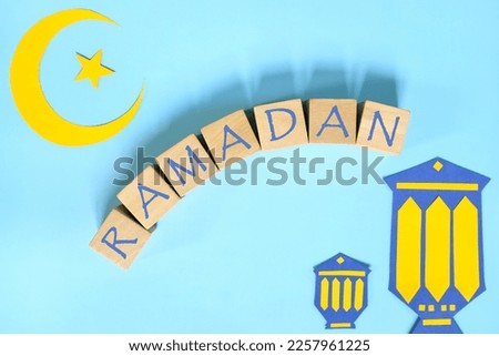 Crescent moon, star and lantern decoration with text on wooden blocks flat lay in blue background. Ramadan Kareem celebration of Islamic holy month.