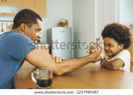 Black African American father competing in arm wrestling with his little boy in kitchen at home