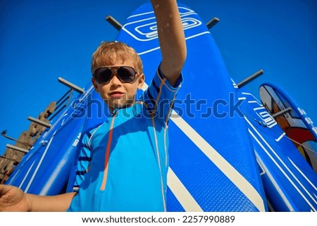 Caucasian child surfer pours sand on the beach in summer on the background of swimming boards. A cool little boy in a surf suit and sunglasses confidently looks at the camera