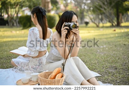 Beautiful hipster Asian woman in casual clothes sits on blanket, enjoying picnic with her friend in the park, taking a picture with her retro camera.