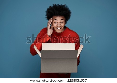 Young happy African American man influencer unpacking delivery box isolated on blue background. Excited customer opening package with birthday gift. Online shopping, sale concept  Royalty-Free Stock Photo #2258031537