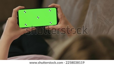 young woman lying on a couch and holding smartphone with horizontal green screen