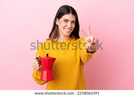 Young Italian woman holding a coffee maker isolated on pink background smiling and showing victory sign