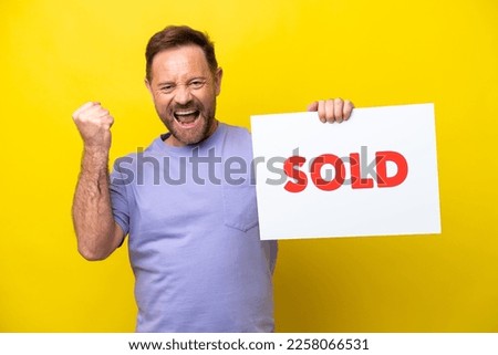 Middle age caucasian man isolated on yellow background holding a placard with text SOLD and celebrating a victory