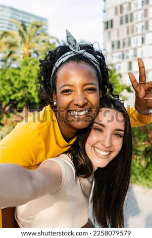 Vertical shot of two multiracial best female friends in casual summer clothes having fun together taking self portrait using phone outdoors.