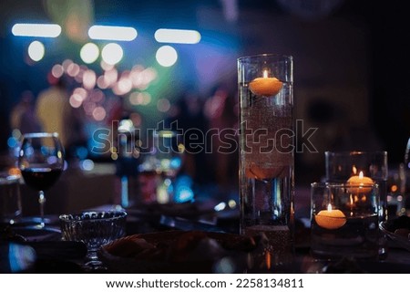 Banquet table decorated with burning candles in glass vases in restaurant hall. In the background party with silhouettes of people dancing on the dance floor with disco lights glowing searchlight. Royalty-Free Stock Photo #2258134811
