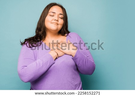 Attractive loving fat woman putting her hand on her heart feeling in love and grateful next to copy space