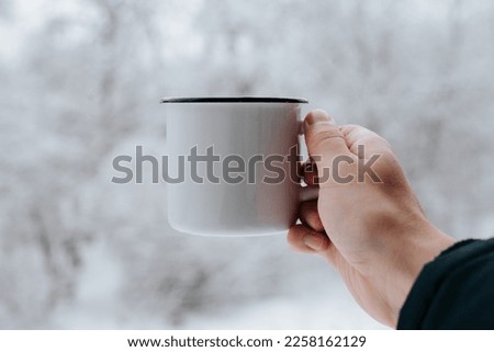 Man holding camping iron white mug against background of snowy forest, close-up.