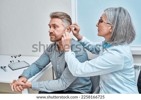 Experienced female doctor fitting Intra-The-Ear hearing aid into patient's ear while consultation in audiology clinic. ITE and ITC hearing aids Royalty-Free Stock Photo #2258166725