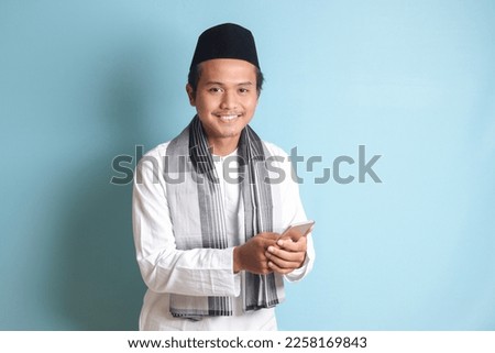 Portrait of young Asian muslim man holding mobile phone with smiling expression on face. Isolated image on blue background