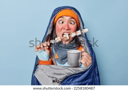 Horizontal shot of funny Asian female hiker crosses eyes has red frozen skin bites delicious roasted marshmallow drinks tea wrapped in sleeping bag isolated over blue background. Female camper