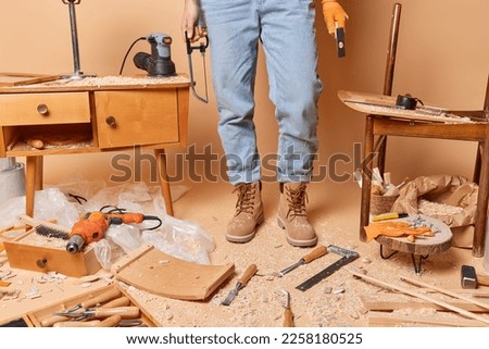 Cropped shot of unrecognizable carpenter in jeans and boots holds instruments poses at messy workshop repairs wooden furniture being professional woodworker surrounded by sawdust. Carpentry concept