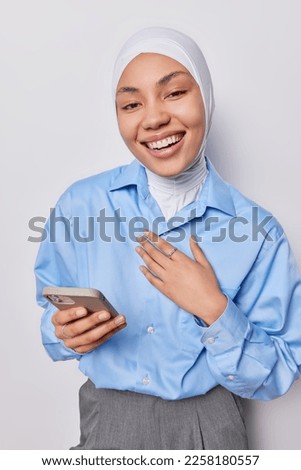 Touched thankful Islamic woman keeps hand on chest holds mobile phone happy to receive congratulations on her birthday wears hijab formal blue shirt and grey trousers isolated over white background