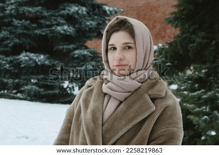 Portrait of a girl in a fur coat and a beige scarf on her head against the background of a green spruce.