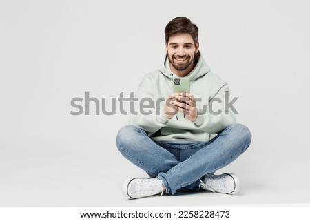 Full body smiling happy cheerful young caucasian man wear mint hoody look camera hold in hand use mobile cell phone isolated on plain solid white background studio portrait. People lifestyle concept