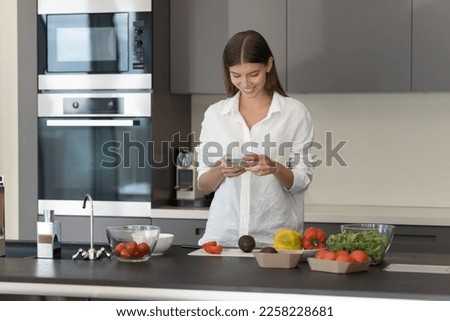 Cheerful millennial girl using mobile phone in kitchen, chatting, typing, consulting cook blog, reading online recipe, preparing salad for dinner, cutting fresh vegetables on table, smiling