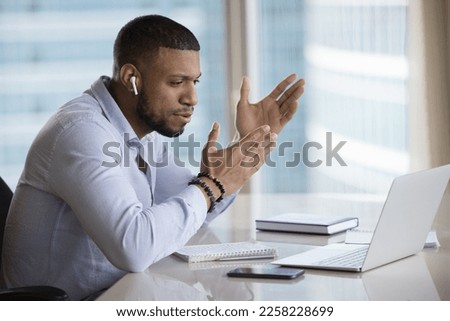 Motivated young African employee with earphone in ear talking on video call in laptop, speaking to customer, giving telephone support. Handsome coach, business blogger speaking at conference Royalty-Free Stock Photo #2258228699
