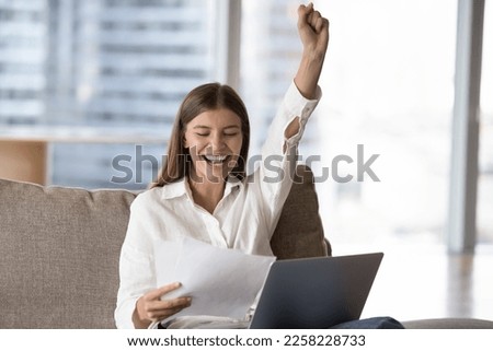 Cheerful excited young freelance employee woman reading legal document, throwing fist up, making winner hand gesture, celebrating good news, study success, career achieve Royalty-Free Stock Photo #2258228733
