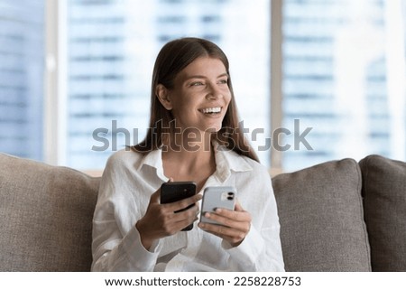 Cheerful pretty young gadget user woman holding two smartphones, sitting on home couch, using wireless connection, data transfer technology, looking away, thinking, smiling, laughing