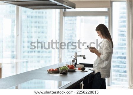 Happy young chef woman using mobile phone while cooking salad, browsing internet, searching for salad recipe, standing in modern home kitchen against large window with city view. Side shot