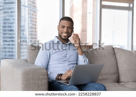 Happy young African businessman home portrait. Cheerful busy handsome millennial freelance entrepreneur talking on mobile phone, using laptop, looking at camera, smiling, sitting on sofa