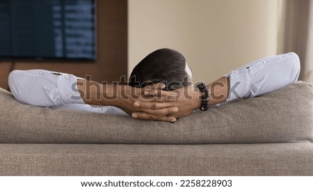 Rear view of head and hands of young Black man resting in sofa, holding nap, looking away, enjoying leisure time, relaxation at home, hotel rom. Close up of arms, couch back