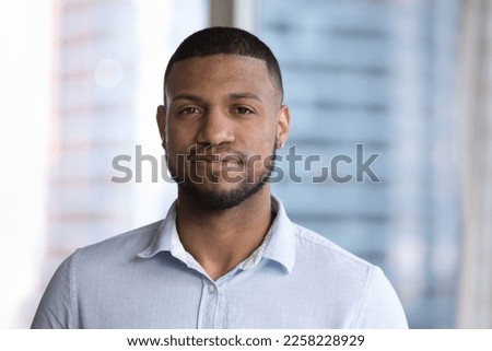 Handsome young African man head shot portrait against blurred big window glass background. Attractive Black male model with stubble, ear piercing wearing pale blue shirt, looking at camera