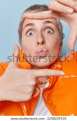 Photo of surprised short haired woman measures angle looks through fingers makes frame pictures something keeps lips rounded creats perfect shot poses indoors against blue background. Body language