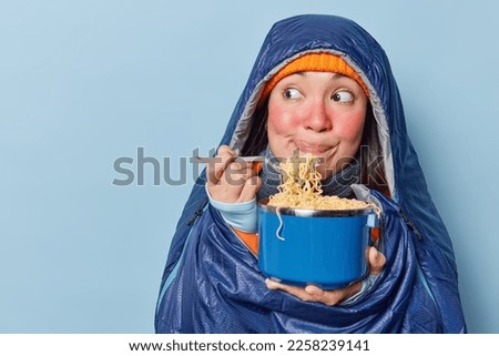 Indoor shot of Asian female hiker eats noodles focused aside licks lips focused aside has red facial skin wrapped in sleeping bag enjoys delicious supper isolated on blue wall copy space for text