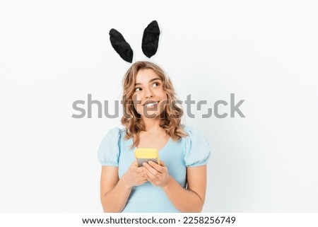 Portrait young woman wearing easter bunny ears holding smartphone smiling looking thoughtfully to the side