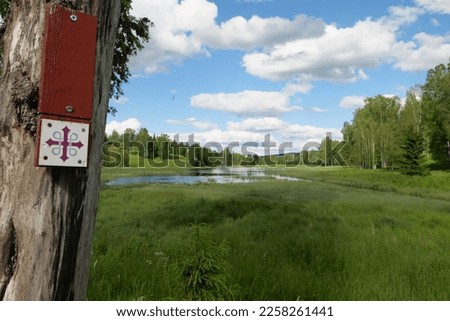 Typical waymarking on the St. Olav's Way, Gudbrandsdalsleden. The pilgrimage route is in Norway goes from Oslo to Trondhein. Here the symbol hangs on a tree trunk at an idyllic pond near Jessheim. Royalty-Free Stock Photo #2258261441