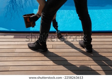 Pool deck terrace renovation. Person applying brown wood protection oil on decking boards with paint brush Royalty-Free Stock Photo #2258263613