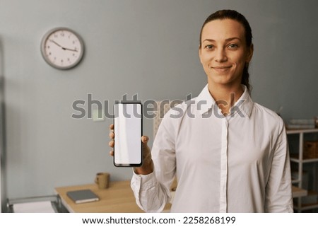 Waist up portrait of smiling young woman holding smartphone with white screen mockup to camera while standing in office setting