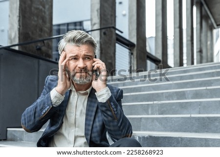 Upset and disappointed mature businessman sitting on stairs of office building outside, man in business suit bankrupt talking on the phone, senior boss investor lost money Royalty-Free Stock Photo #2258268923