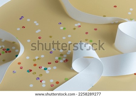 White fabric grosgrain ribbon curling around a pale gold background with colorful confetti dots. High angle view, shallow depth of field, Concept of party, special occasion, surprise.