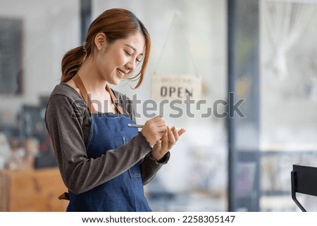 Shot of smiling young cafe show owner Asian woman standing with arms crossed and Open sign on the glass door. Portrait of asian tan woman barista cafe SME entrepreneur seller business concept.