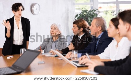 Concentrated young adult old office workers sitting around the table and looking at screen while Latin female using projector remote control in office