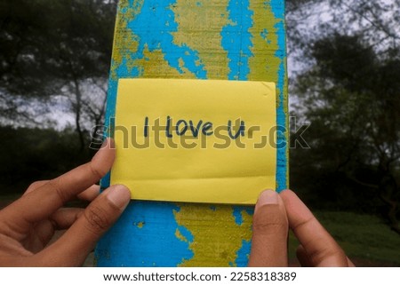 Aesthetic of a hand holding a yellow sticky note saying "I Love U" on a wooden background. The beautiful Wallpaper message hanging on the wall to the loved one