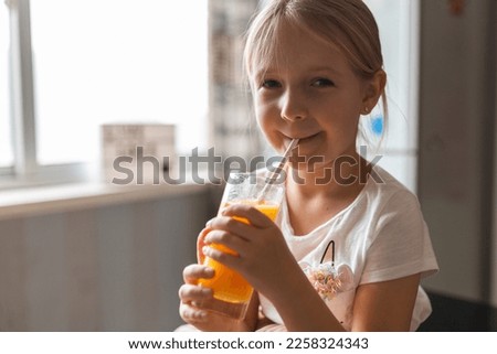 Cute little caucasian girl with blonde hair sitting on the kitchen table and drinking fresh mango juice. Healthy lifestyle concept. Kid 6 years old at home