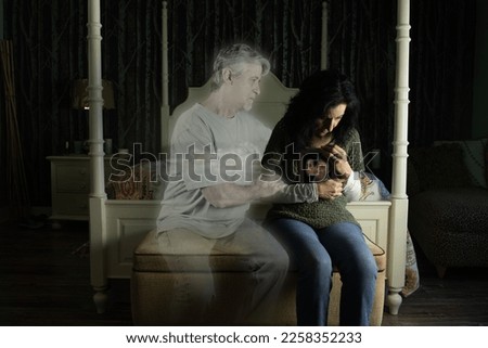 A sad woman clutching a photo of herself and her dead husband is being consoled by his ghost as she sits by her bed crying in her dimly lit bedroom. Life after death, heaven and the afterlife. Royalty-Free Stock Photo #2258352233
