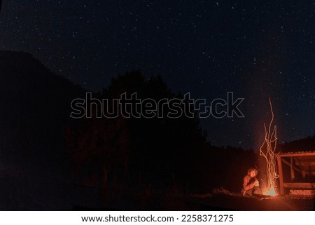 Lonely tourist girl sitting by the flames with sparks of a fire at night under the starry sky next to the edge of the gazebo and the mountain.