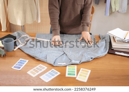 Woman designer entrepreneur using measure tape to measuring chest size of shirt and checking detail while working with design new collection in home workshop.