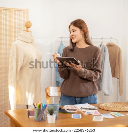 Woman designer entrepreneur checking detail of sweater in mannequin and writing data in tablet while working to sketching design new collection for winter in home workshop.