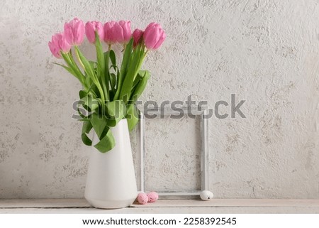 Vase with beautiful tulip flowers, Easter eggs and empty picture frame on table near light wall